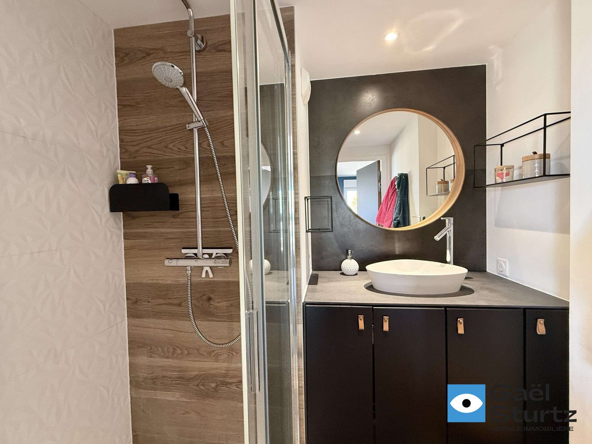 Salle d’eau avec douche vitrée, vasque et miroir rond.